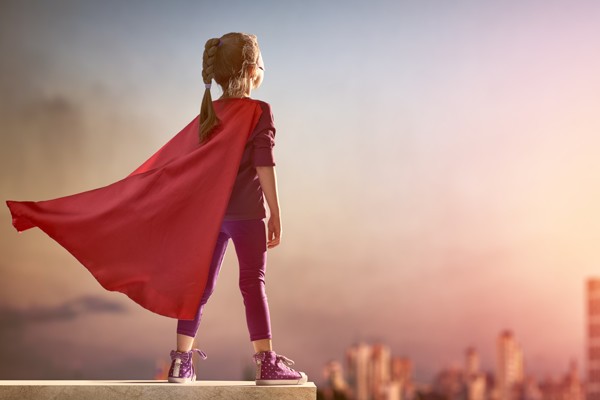 little girl in a billowing red cape with her back to us on top of a wall overlooking a city