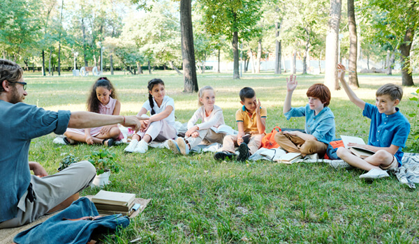 A group of six elementary school students sit in a semicircle on the grass. One has his hand raised and the teacher opposite points to him.