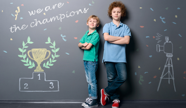 Two young boys standing in front of a blackboard wall, on the wall is written 'We are the champions'