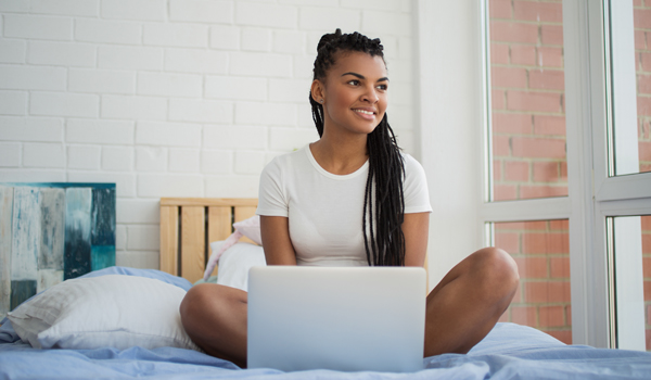 African writer sitting on her bed and working on a laptop while gazing thoughtfully out the window.