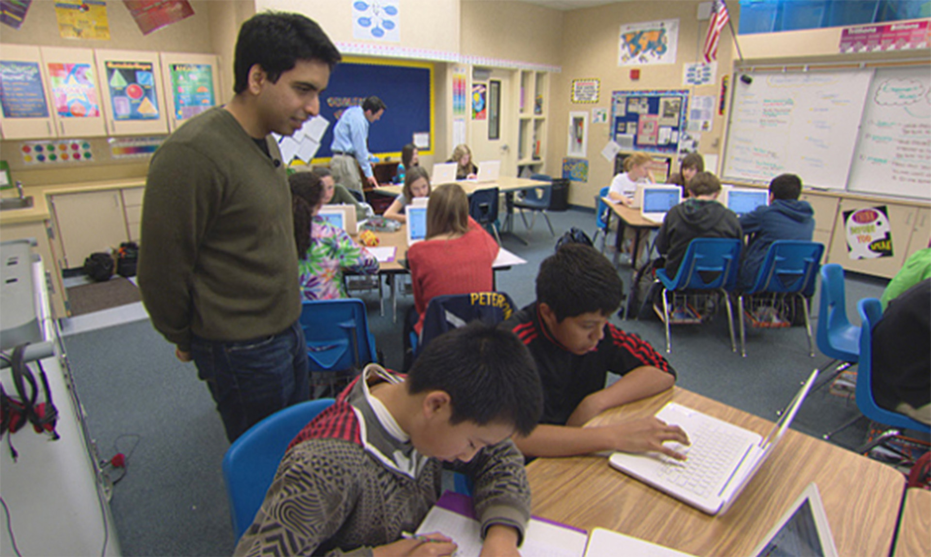 Students in a classroom work on paper and laptop while two teachers observe them.