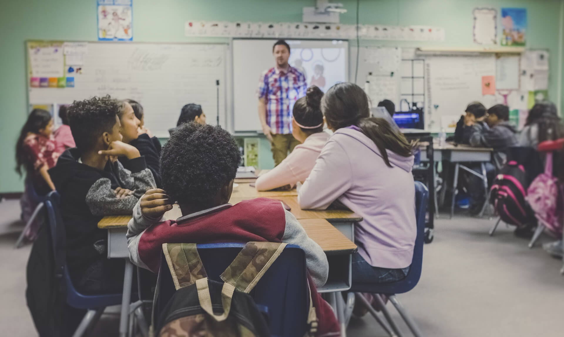 A group of high school students listen to a teacher at the front of the class.