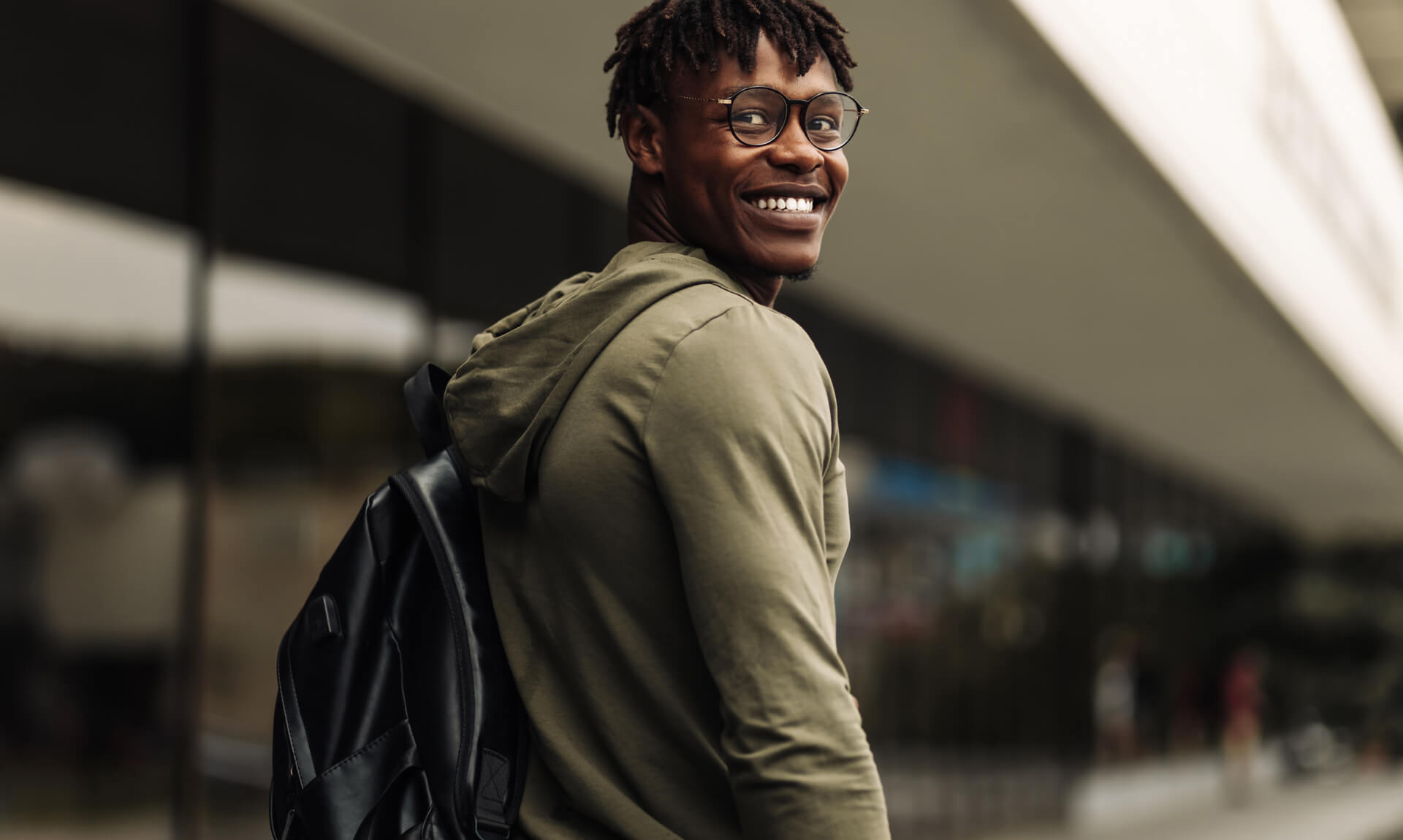 Happy successful african student, wearing glasses and with a black backpack, standing on the steps and smiling