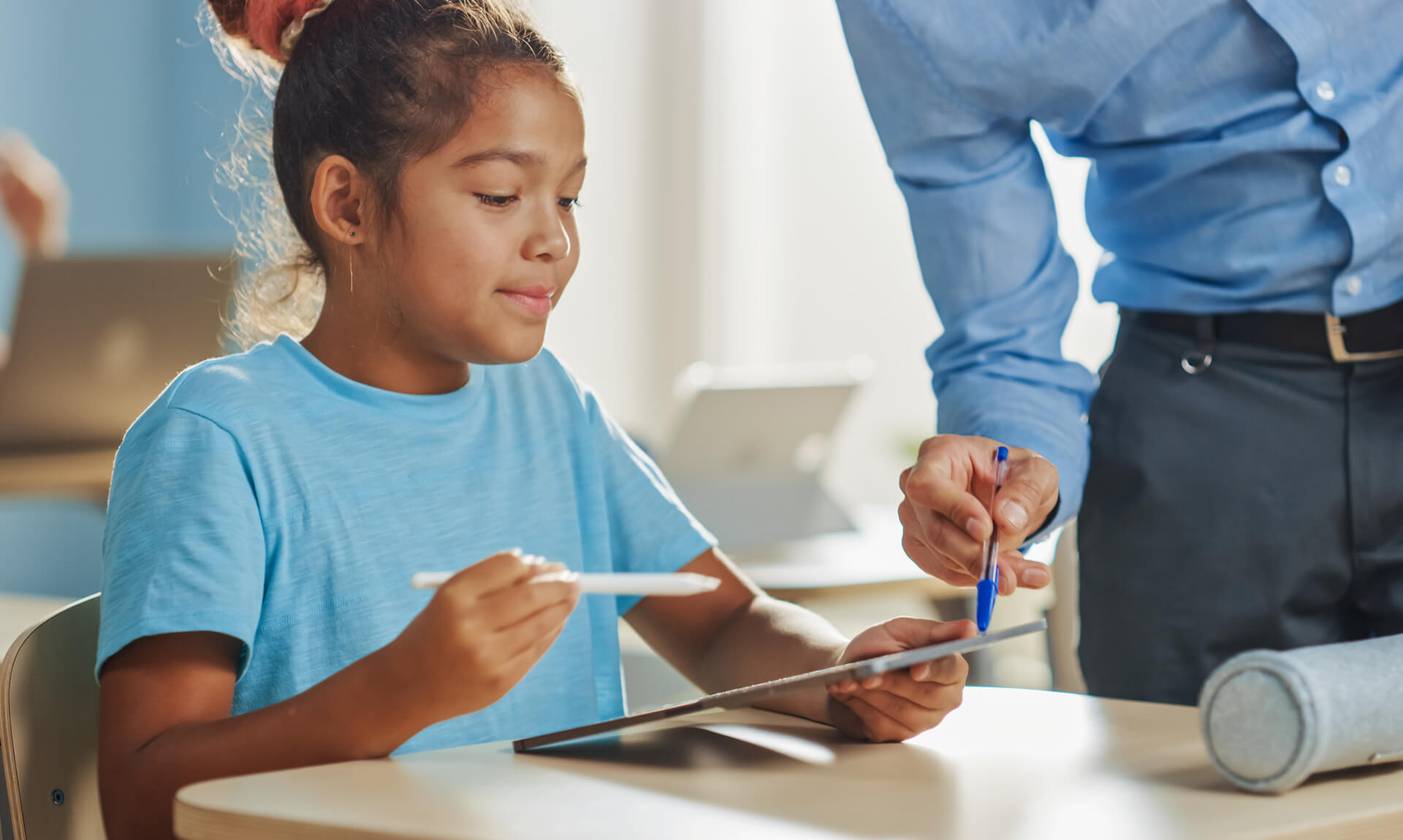 Girl uses digital tablet while teacher helps her by explaining the assignment.