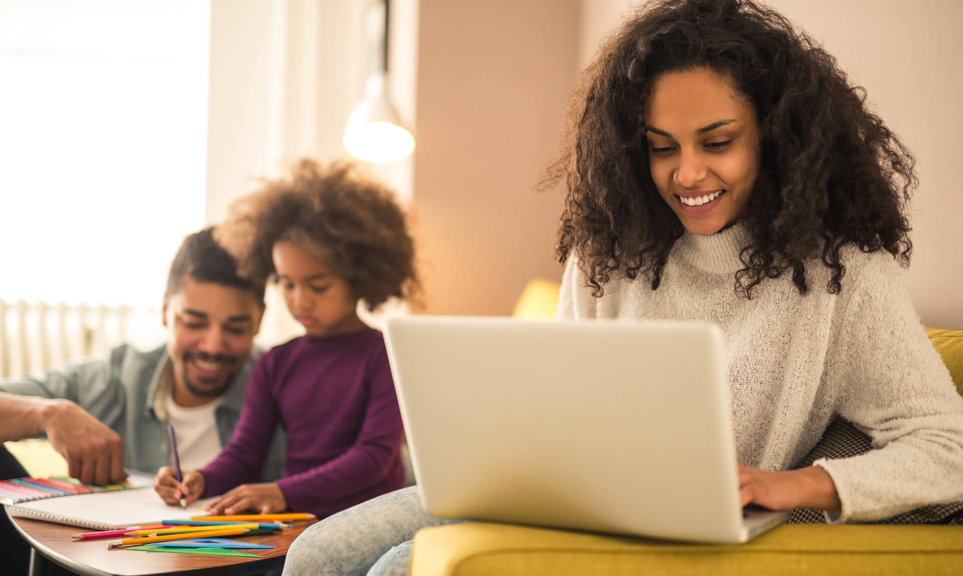 Mom works on computer while dad watches child write in notebook in background.