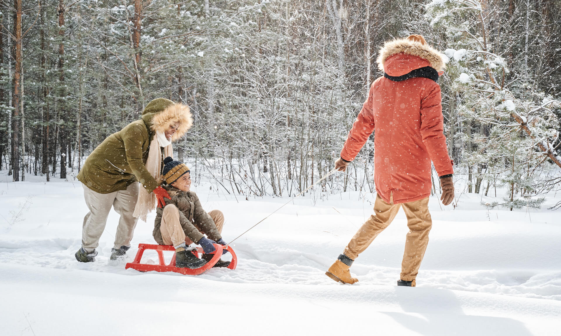 woman pushing sledge with her happy little son while man in winterwear pulling it during walk in forest on winter day