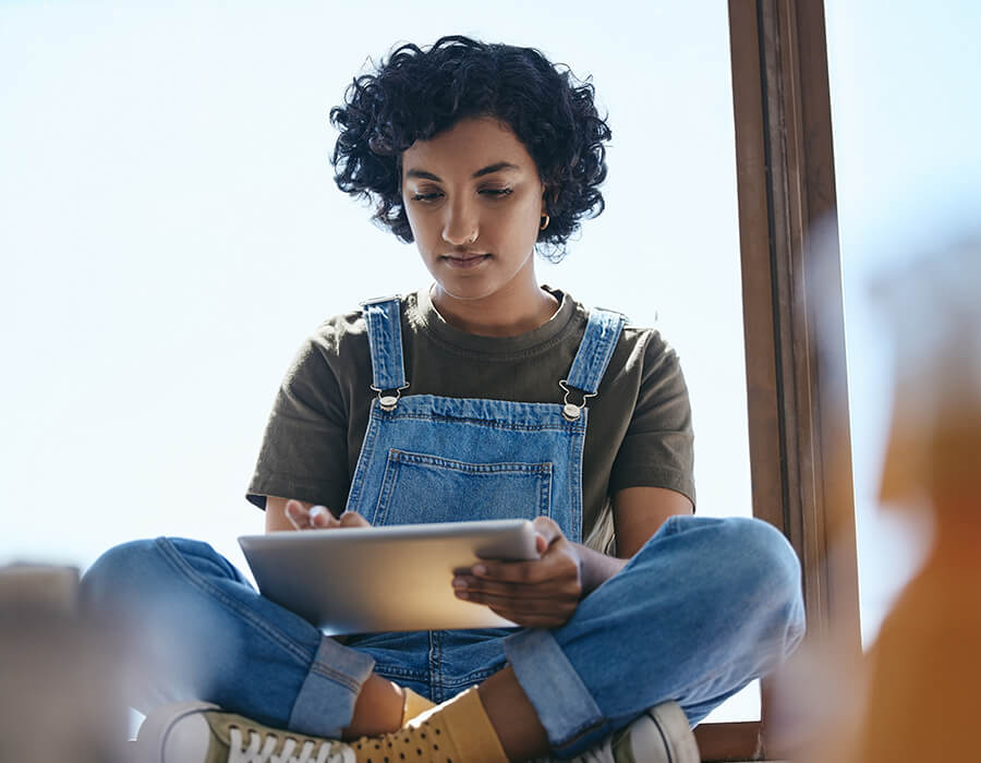 Join our email list Young woman with short curly hair sits cross-legged by a window, focused on using a tablet. She wears denim overalls, a green T-shirt, and yellow socks, with natural light illuminating her face.
