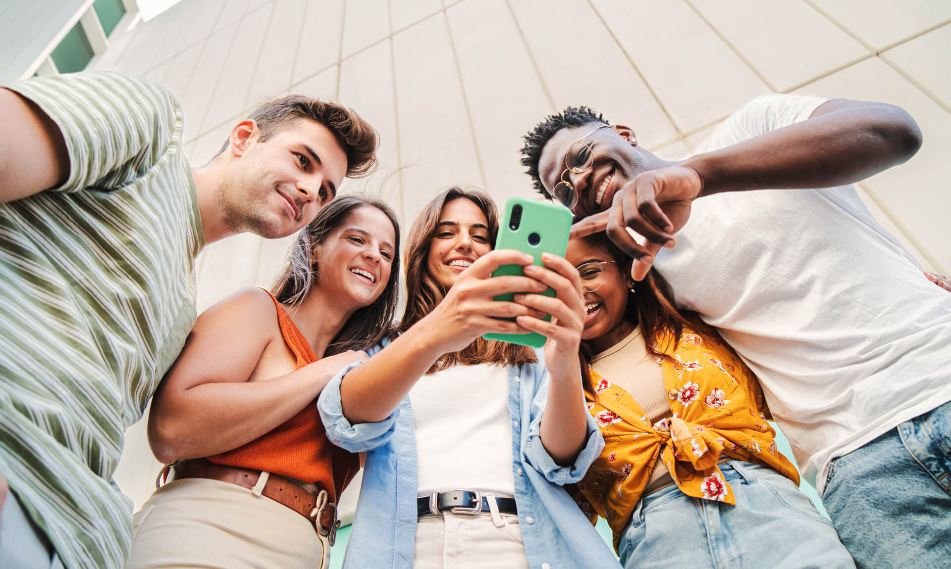 A diverse group of young adults stand in a circle outdoors, smiling and looking down together at a smartphone.