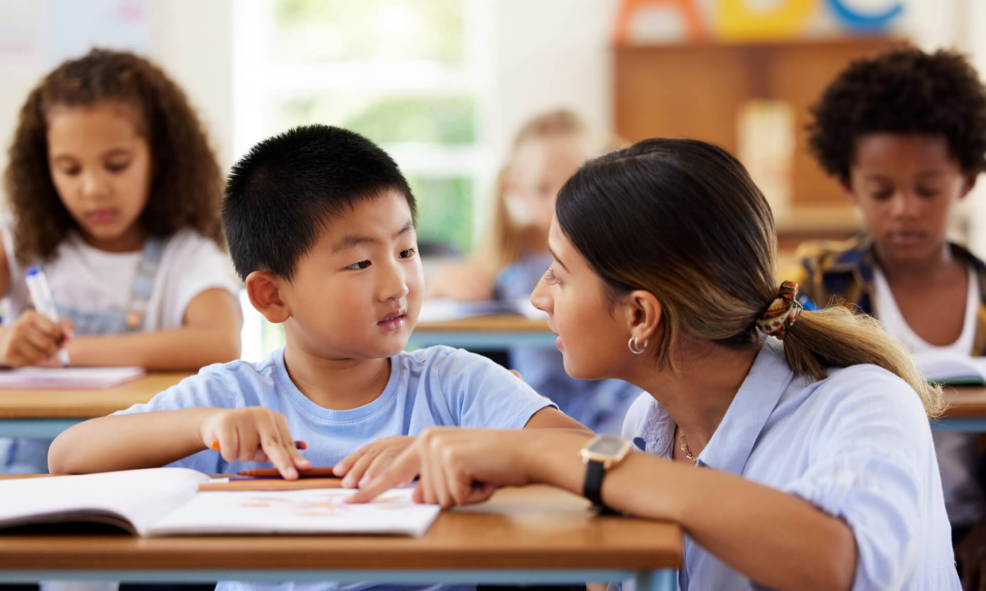 Teacher provides one-on-one reading support to a student in an elementary classroom while other children work independently in the background.