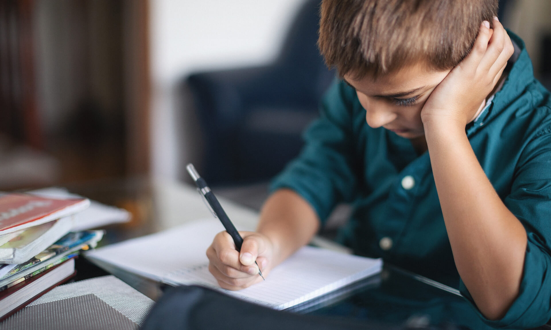 Child leaning on hand while writing in a notebook at a desk, appearing focused and slightly frustrated, with books stacked nearby.
