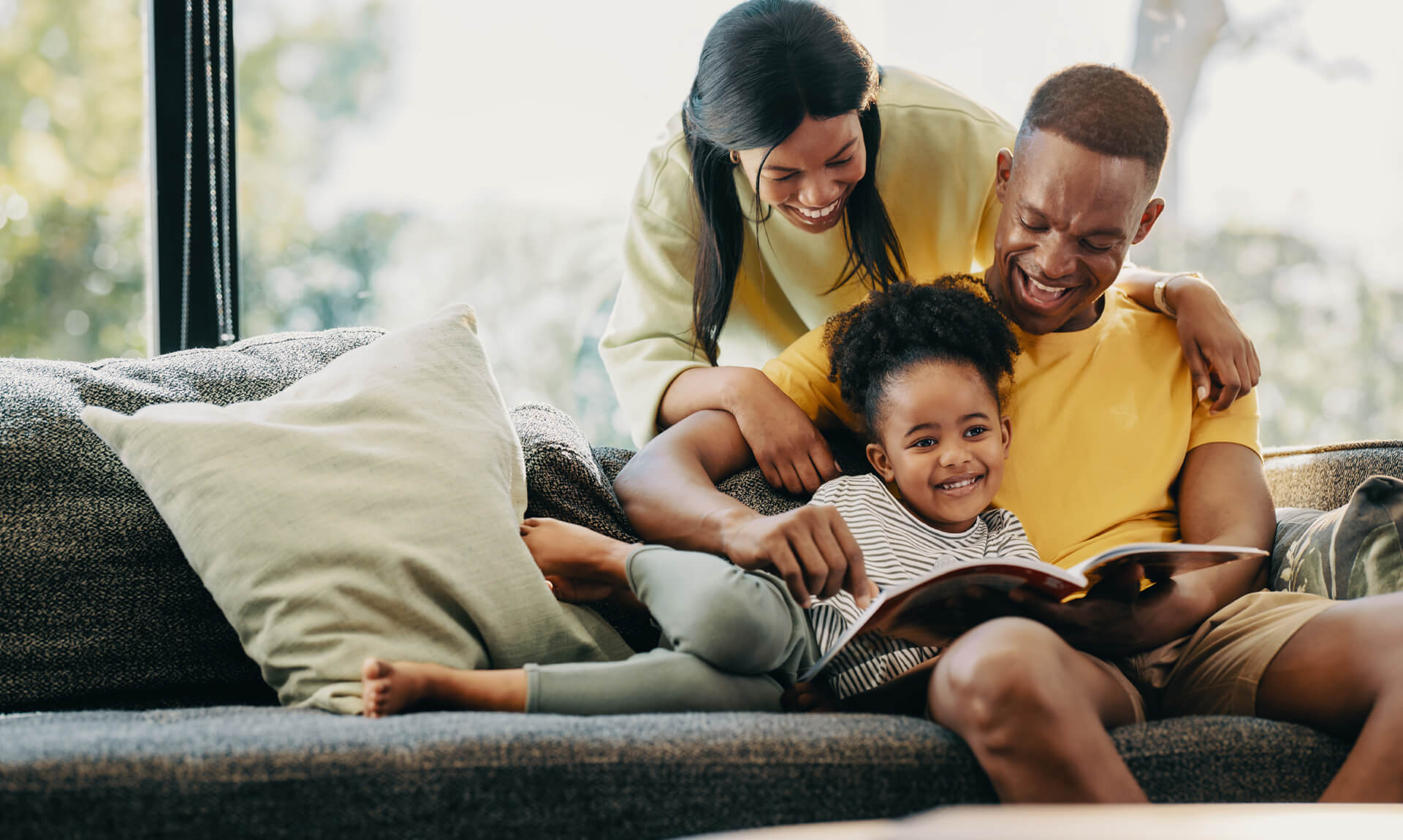 family laughs together on couch while reading a book with daughter