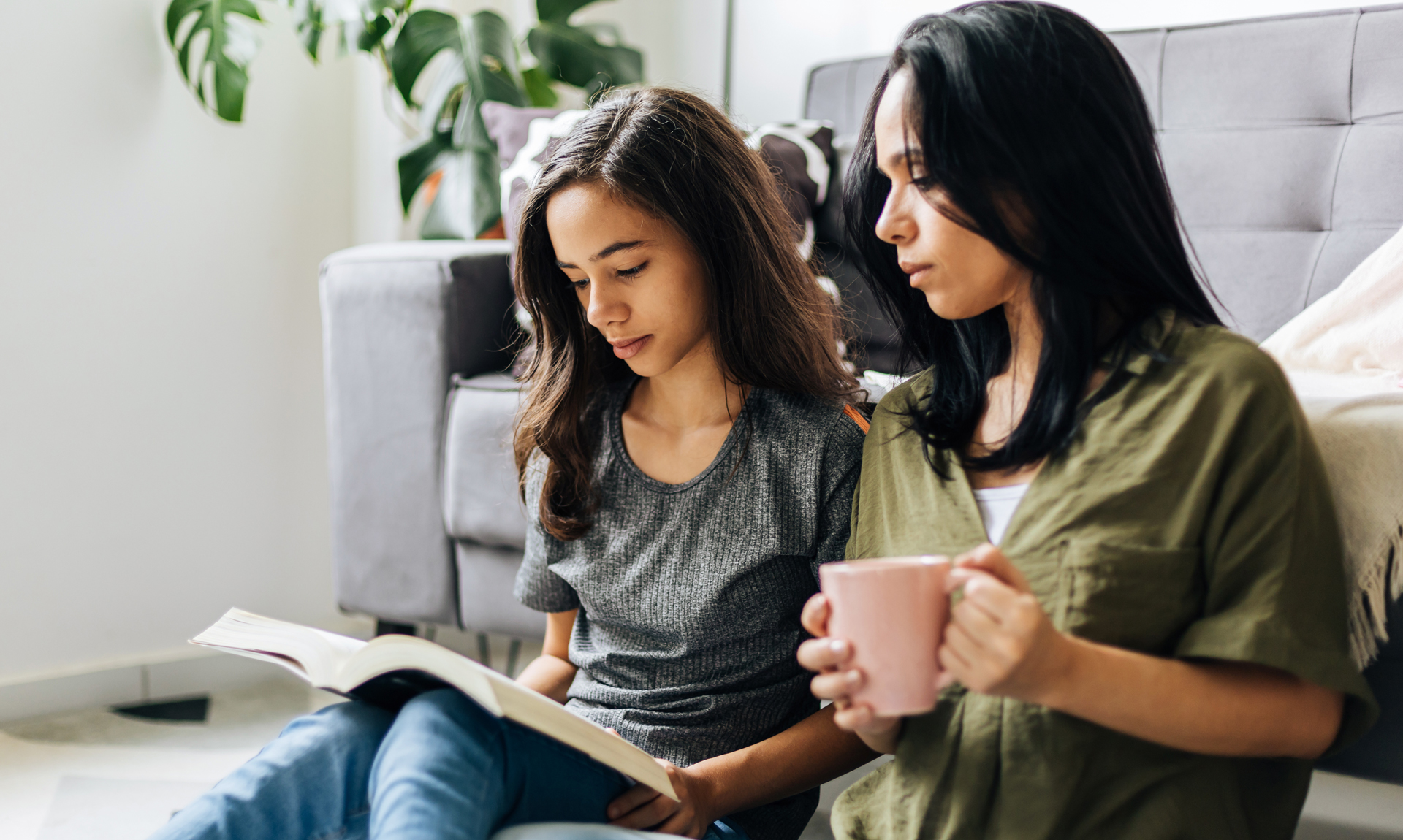 woman drinks coffee while watching daughter read a book
